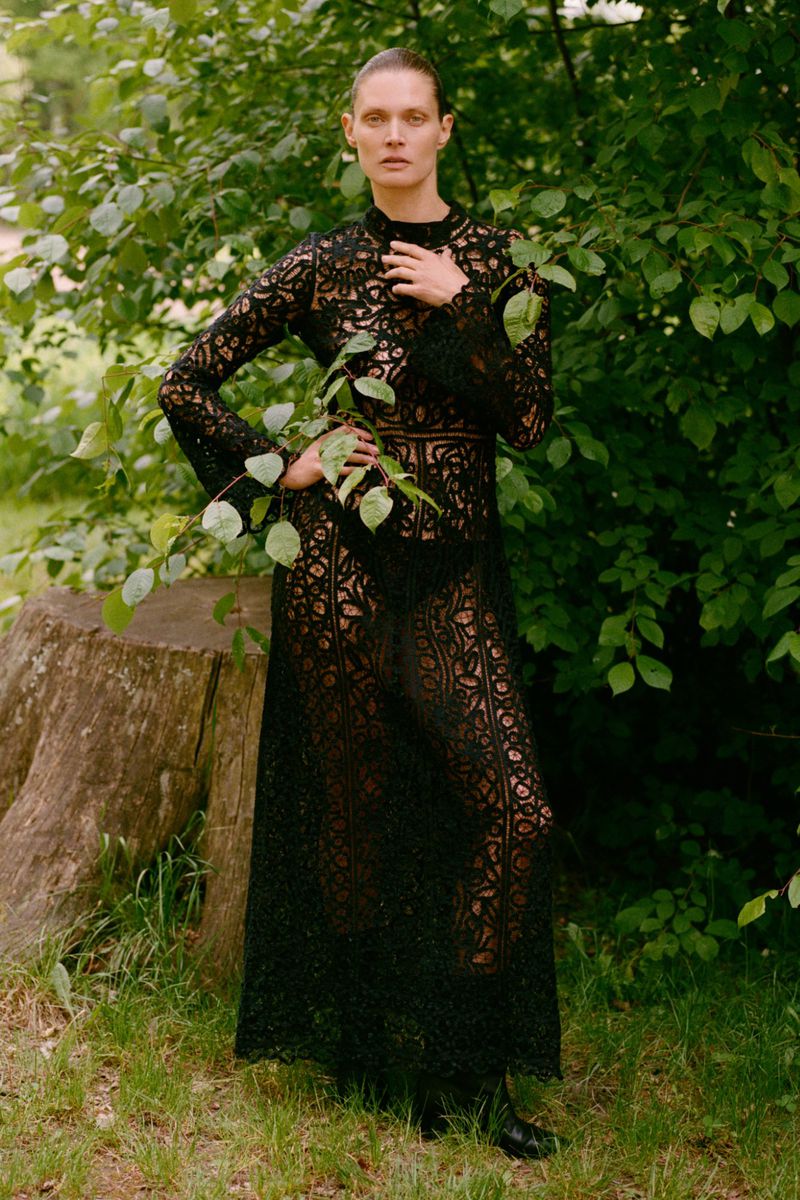 An adult woman with fair skin poses in a full-length black lace dress outdoors, surrounded by greenery and a tree stump in the background.