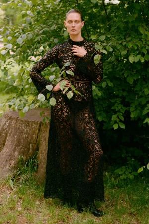 An adult woman with fair skin poses in a full-length black lace dress outdoors, surrounded by greenery and a tree stump in the background.