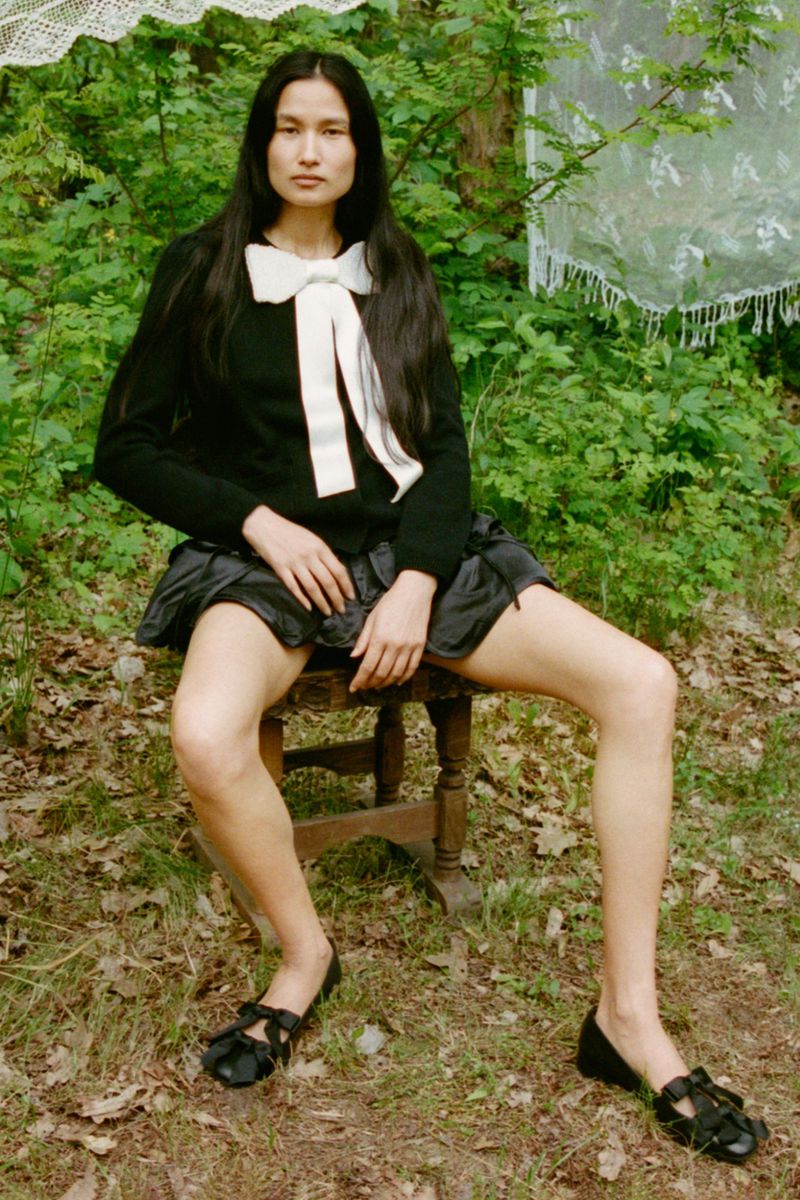 A woman with long dark hair sits on a wooden stool outdoors in a black sweater, black skirt, and Mary Jane shoes in a woodland setting.