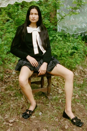A woman with long dark hair sits on a wooden stool outdoors in a black sweater, black skirt, and Mary Jane shoes in a woodland setting.