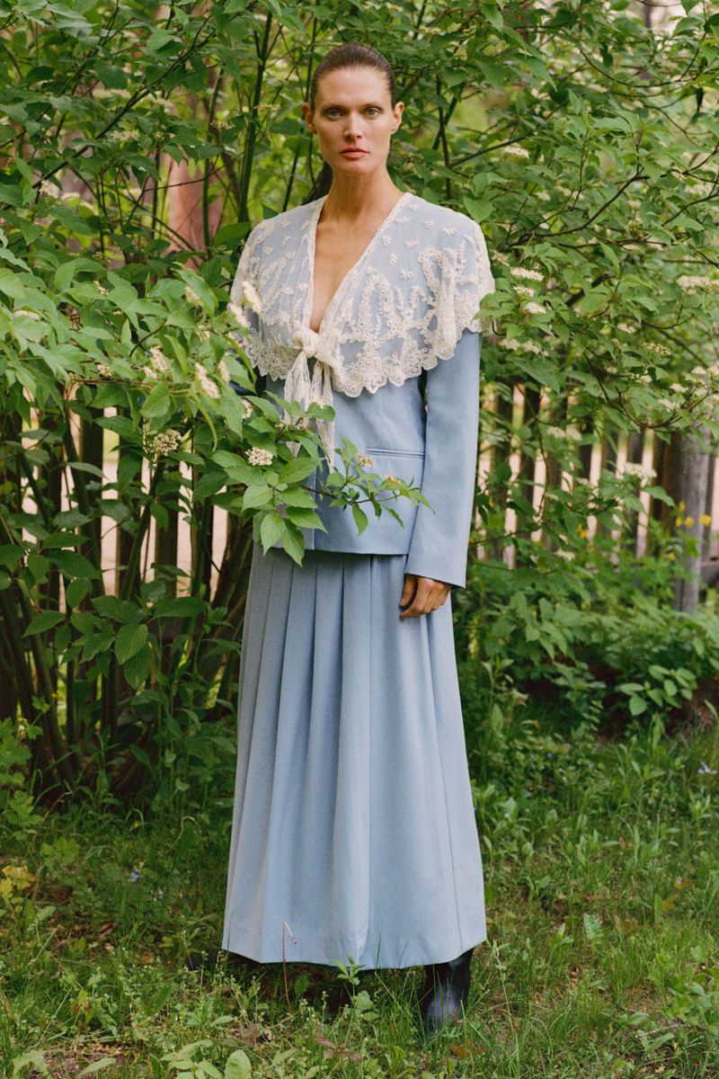 Adult female model in a blue skirt suit with lace collar, standing outside among green leafy plants and trees. Fashion editorial.