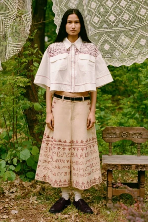 Full-length shot of a woman in a white cropped shirt and a beige skirt with embroidery standing in a wooded area with a lace backdrop.