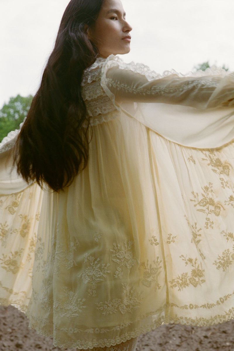 A young woman with long brown hair poses in a flowing white dress with floral embroidery outdoors against a blurred natural background.