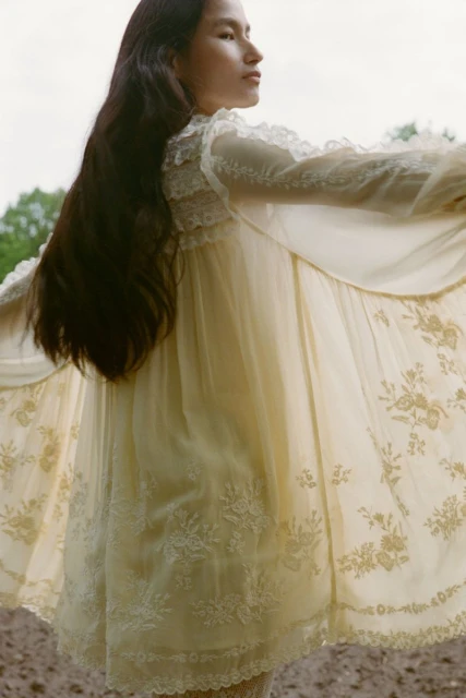 A young woman with long brown hair poses in a flowing white dress with floral embroidery outdoors against a blurred natural background.