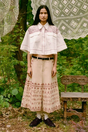 Model Varsha Thapa stands outdoors wearing an embroidered cropped white shirt and matching wide-leg culottes beside a wooden chair.