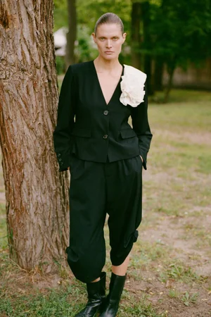 Malgosia Bela standing beside a tree wearing a black tailored jacket with a white flower corsage, cropped trousers, and black boots.