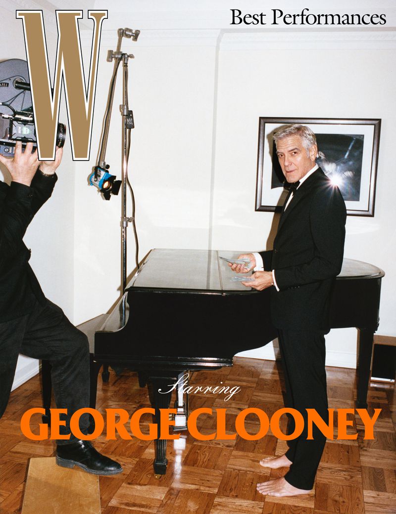 Magazine cover featuring George Clooney in a suit standing next to a piano, photographed indoors with a camera in the foreground.