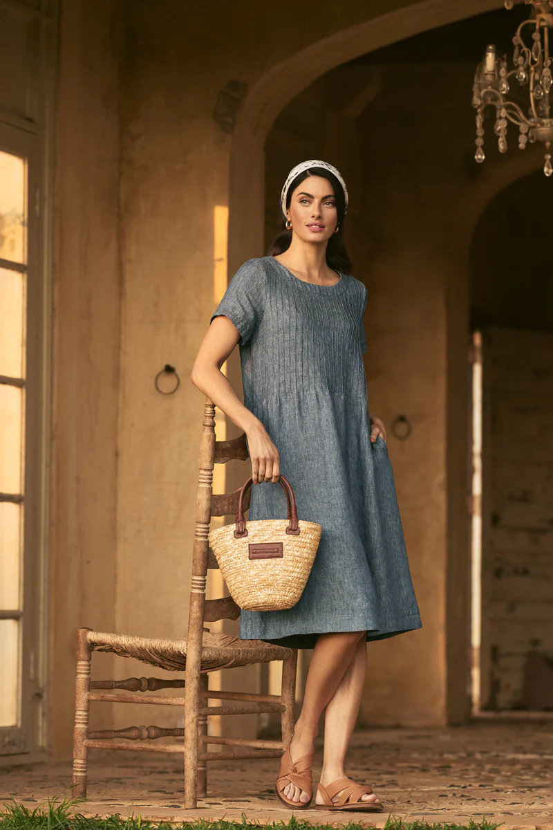 A woman in a blue dress stands in a doorway holding a straw purse. She is standing in front of a wooden chair. 