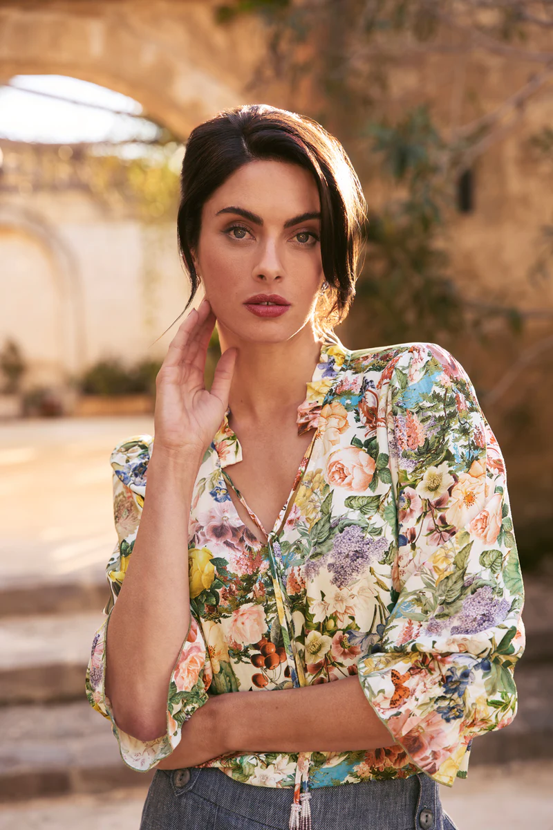 Close-up portrait of a brunette woman in a floral top, posing outdoors in front of an arched stone building on a sunny day.
