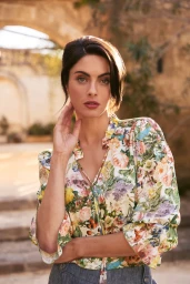 Woman wearing a floral print blouse touching her neck with a stone courtyard and archway in the background.