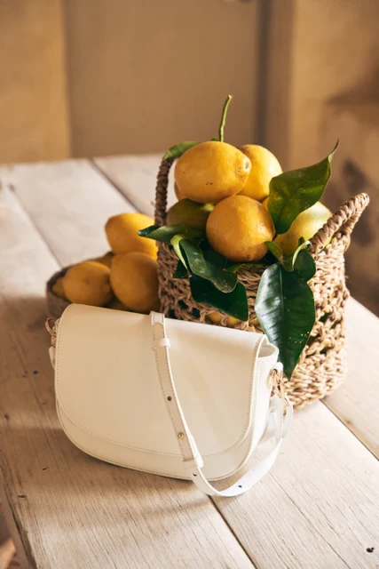 Still life shot of a woven basket full of lemons, with leaves, and a white leather handbag on a rustic wooden surface.