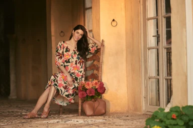 Woman in floral print dress seated on a wooden chair holding her hair against a warm building exterior background