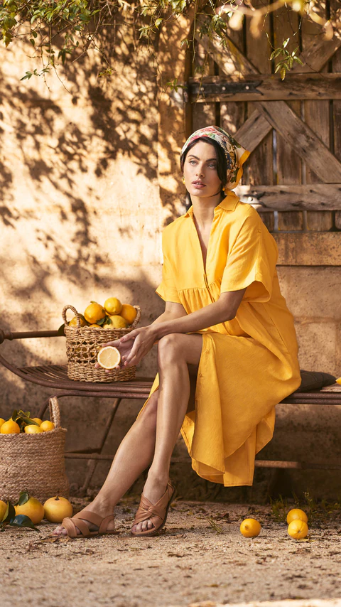Adult woman in a yellow dress sits on a bench next to lemons in baskets near a wooden wall with tree shadows overhead.