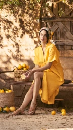 Model wearing yellow dress and patterned headscarf sitting on a bench holding half a lemon outdoors with baskets of lemons.