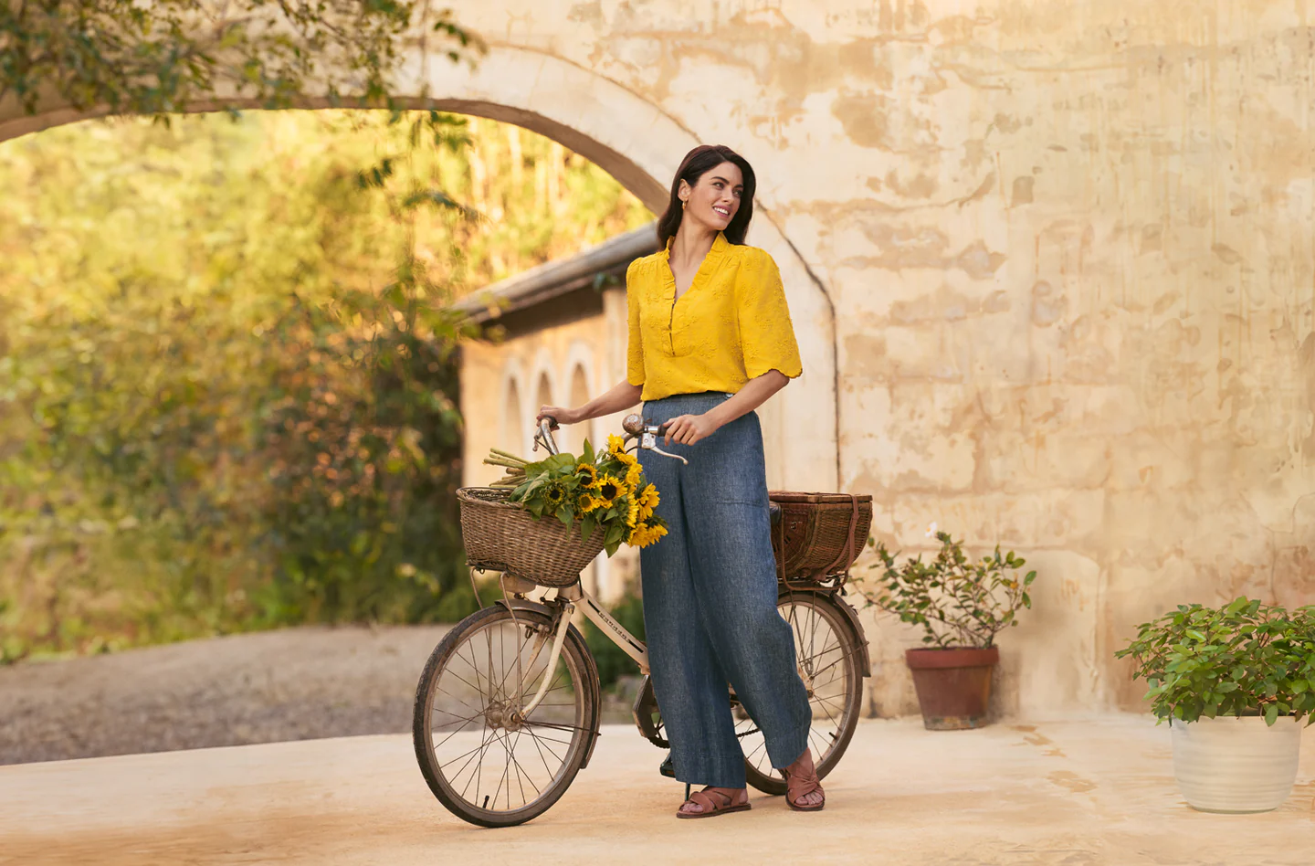 Woman in yellow blouse with bicycle and sunflowers near stone archway and building, creating a charming and sunlit scene.