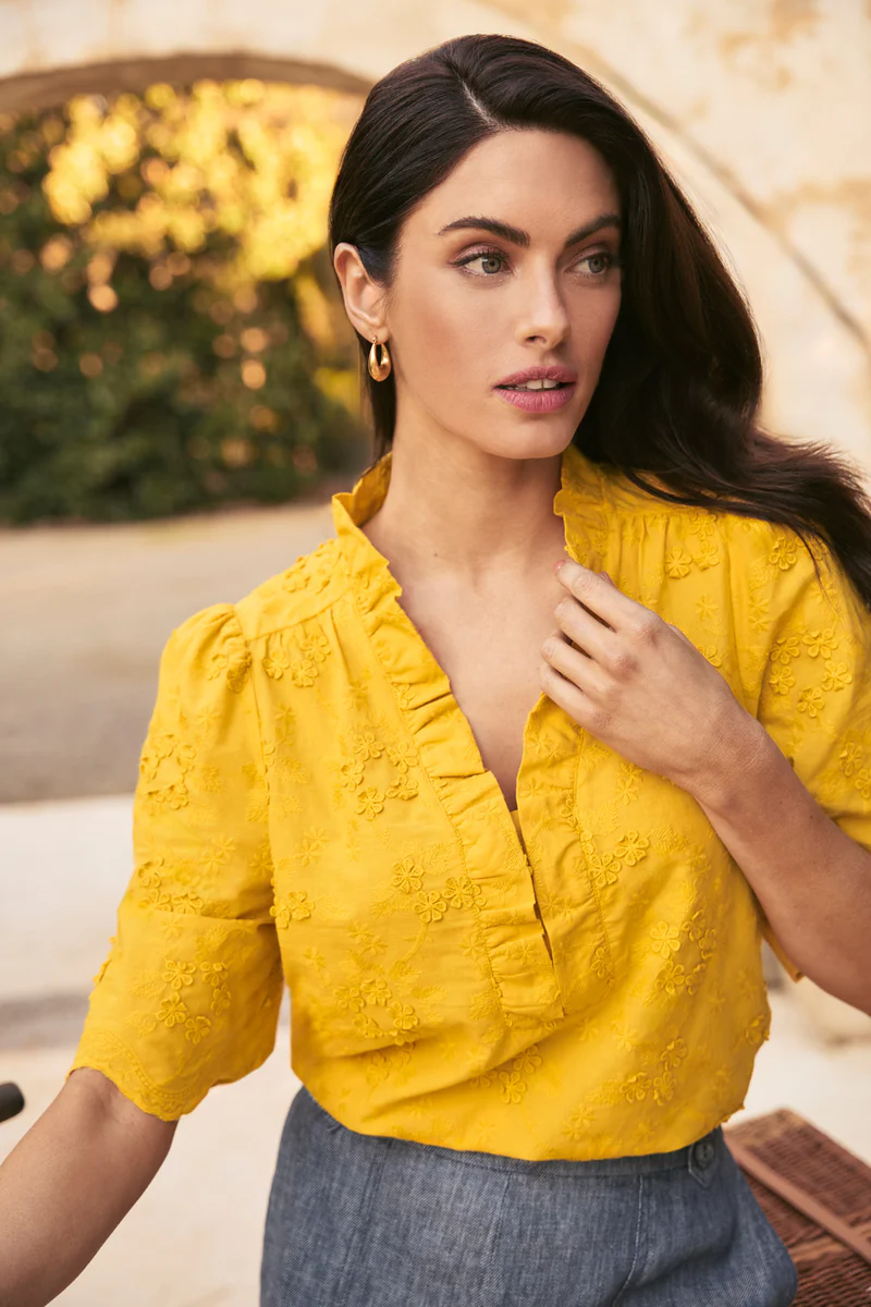 Woman in a yellow embroidered top poses outdoors near a stone structure with greenery; medium shot, fashion, beauty, and lifestyle.