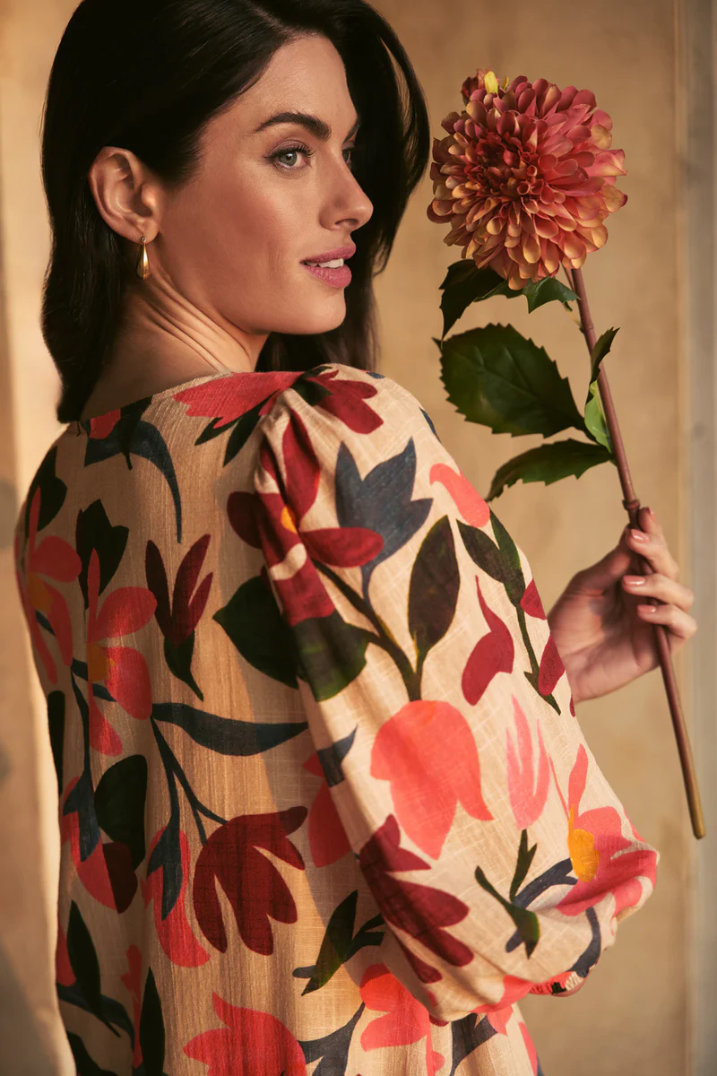 Portrait of a brunette woman in a floral print dress, holding a large dahlia flower against a neutral backdrop in soft studio lighting.