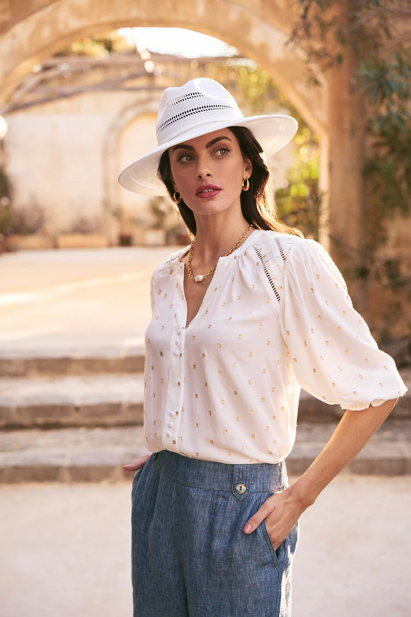 Fashionable woman in blouse and trousers poses in sunlit courtyard, wearing a white hat, gold jewelry, and with hand in pocket.