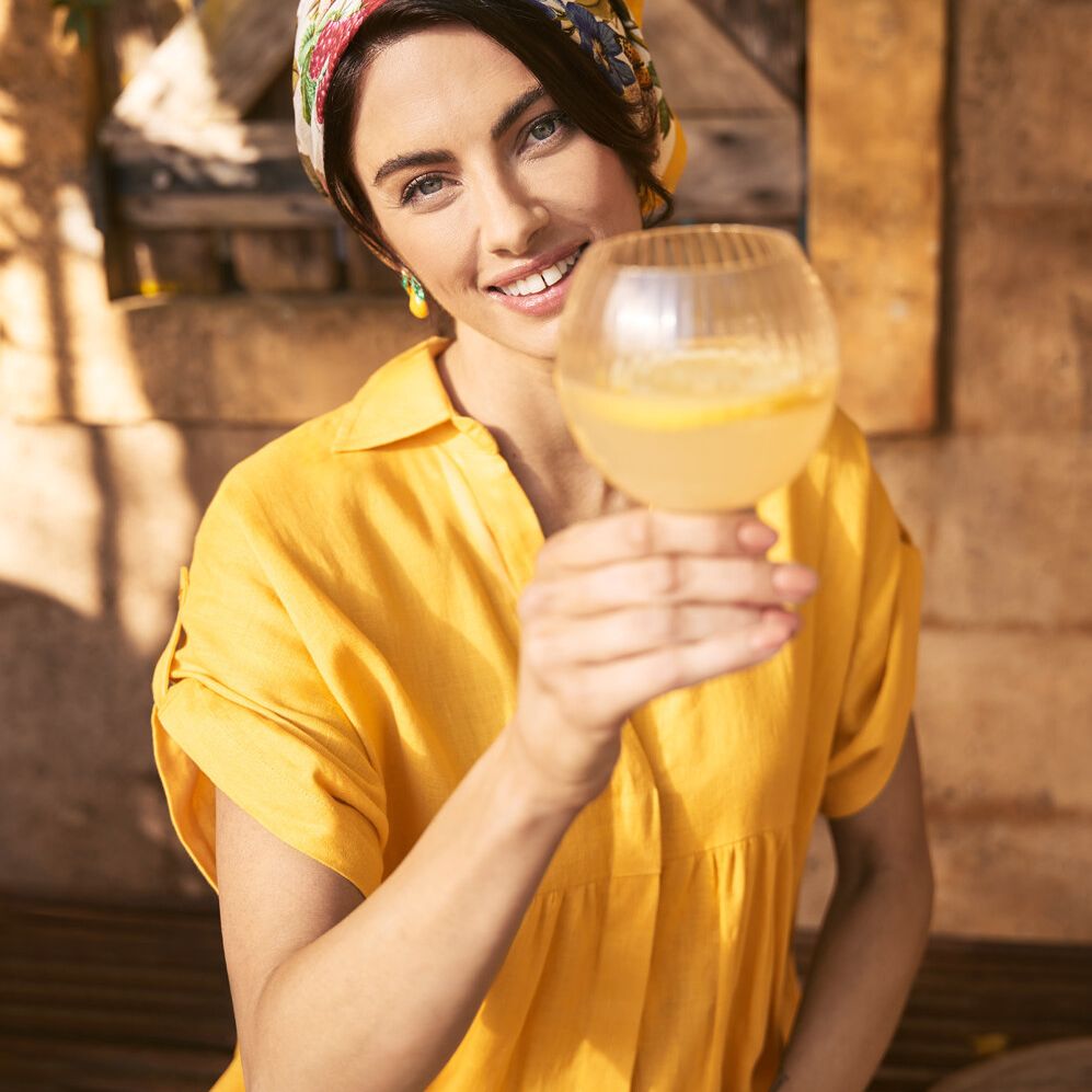 A smiling woman in a yellow dress and floral headscarf raises a stemmed glass filled with a light-colored beverage, outdoors.