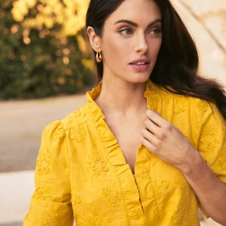 Brunette woman in yellow embroidered top with ruffled collar gazes off-camera, accessorized with gold hoop earrings, outdoor setting.