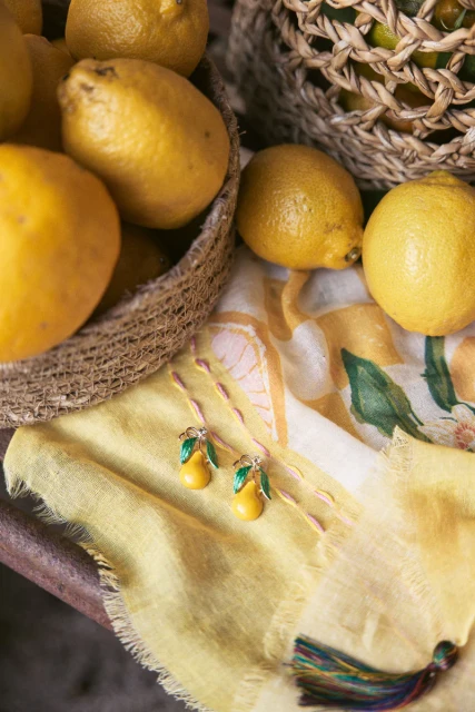 Close-up of lemon-themed earrings, yellow scarf, lemons in baskets. Jewellery product shot for fashion or lifestyle content, studio setting.