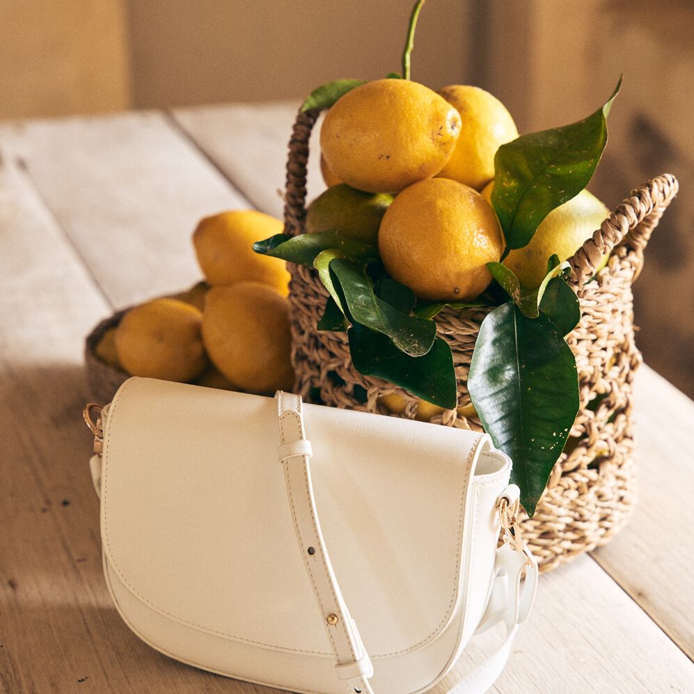 Still life featuring a white crossbody purse on a wooden table next to a woven basket filled with lemons and green leaves.