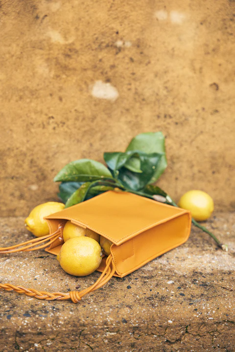 Close-up of a yellow leather handbag filled with lemons and leaves, set against a mottled, neutral-toned wall. Bright, natural light.