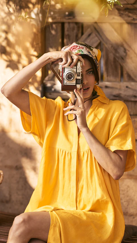Woman in yellow dress captures the moment with vintage camera. Outdoor portrait with warm lighting and headscarf. Editorial fashion photography.