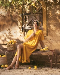 Woman in a yellow midi dress sitting on a rustic bench next to baskets of fresh lemons outdoors