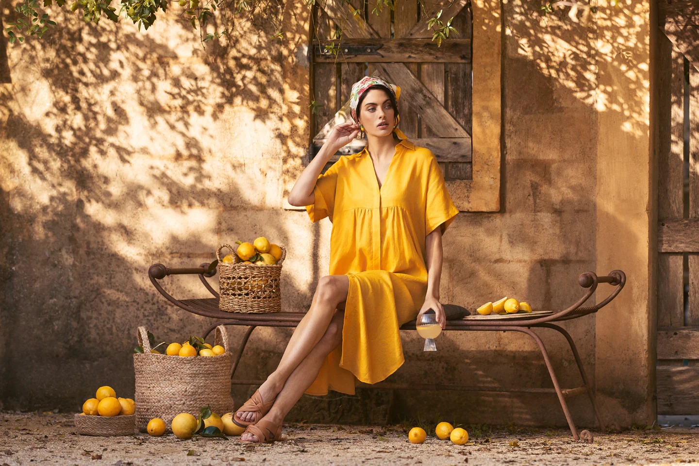 Woman in a yellow dress sits on an iron bench with lemons, wearing a headscarf in an outdoor setting with dappled light.