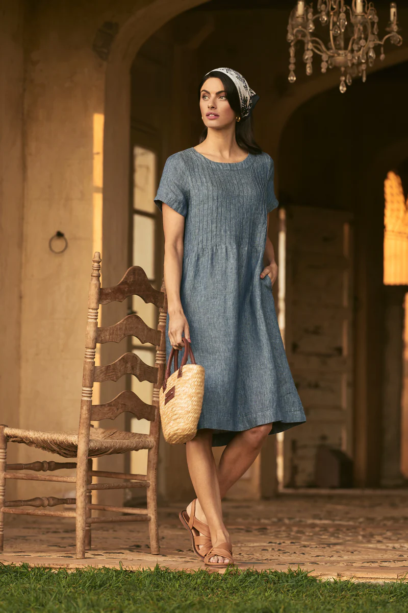 A woman in a blue dress and headscarf carries a straw purse while standing in front of an arched building with a chandelier.