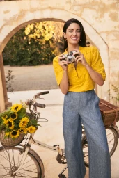 Woman in yellow top and blue trousers holding a vintage camera near a bicycle with sunflowers