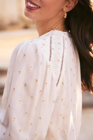 Elegant woman with dark hair, wearing a patterned white top and gold jewelry, smiles softly in a bright, outdoor setting.