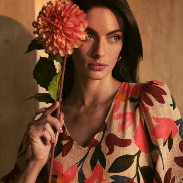 Close-up of a woman with dark hair holding an orange dahlia flower, wearing a printed dress, against a neutral, subtly textured wall.