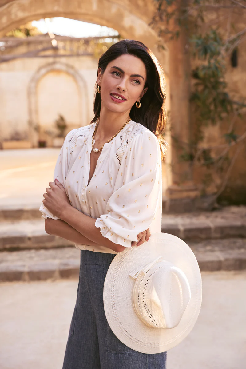 A fair-skinned woman poses outdoors in an off-white blouse and blue pants holding a straw hat with an arched stone structure in the background.