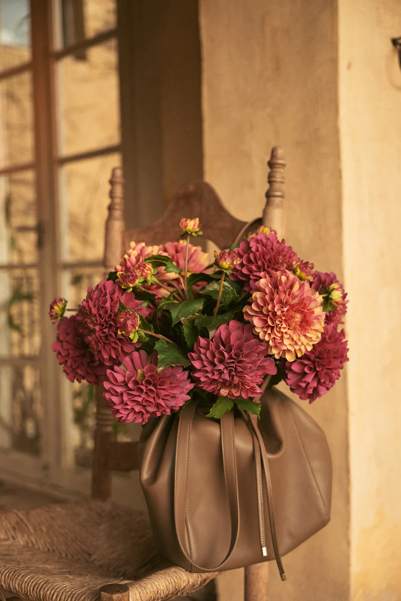Burgundy dahlia flowers arranged in a taupe leather drawstring handbag placed on a rustic woven chair against a tan wall indoors.