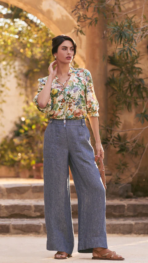 A woman in a floral blouse and wide-leg denim pants stands in an outdoor courtyard with an archway and stone steps, bathed in sunlight.