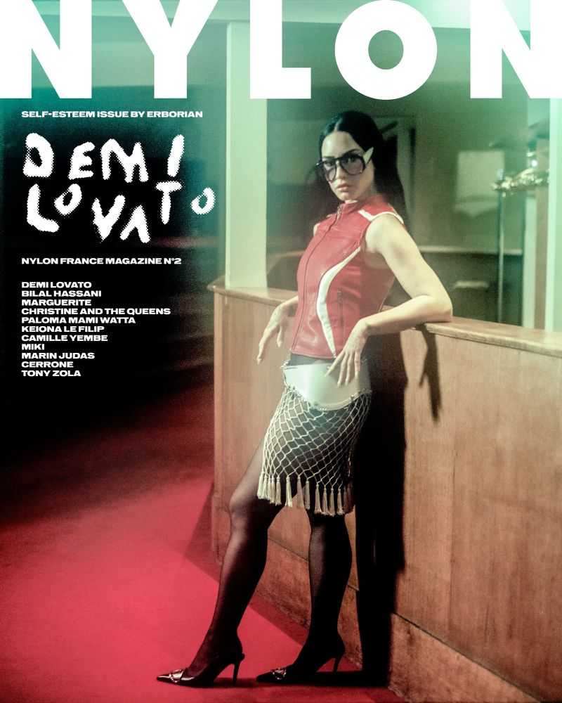 Demi Lovato poses for a magazine cover in a red vest, patterned skirt, and heels, set against a backdrop featuring a doorway and text.