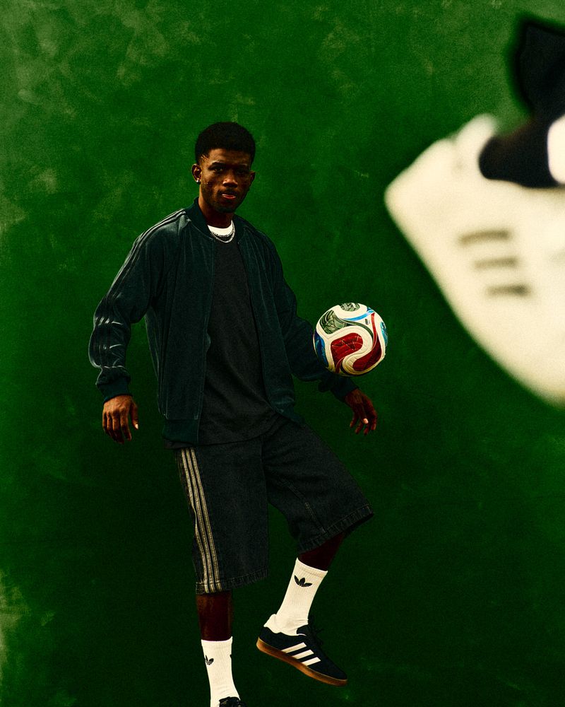 An African American man balances a soccer ball in a fashion-style full shot against a green background, showcasing athletic wear and casual style.