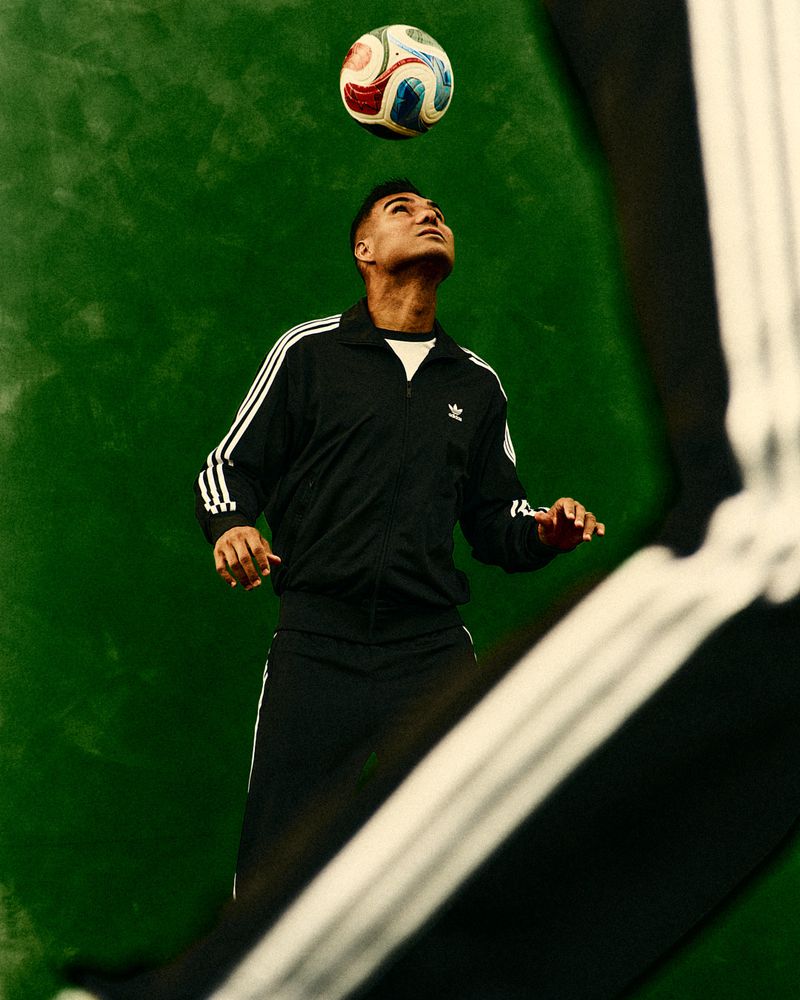 Adult male in black Adidas tracksuit balances a soccer ball on his head against a solid green backdrop in an eye-level medium shot.