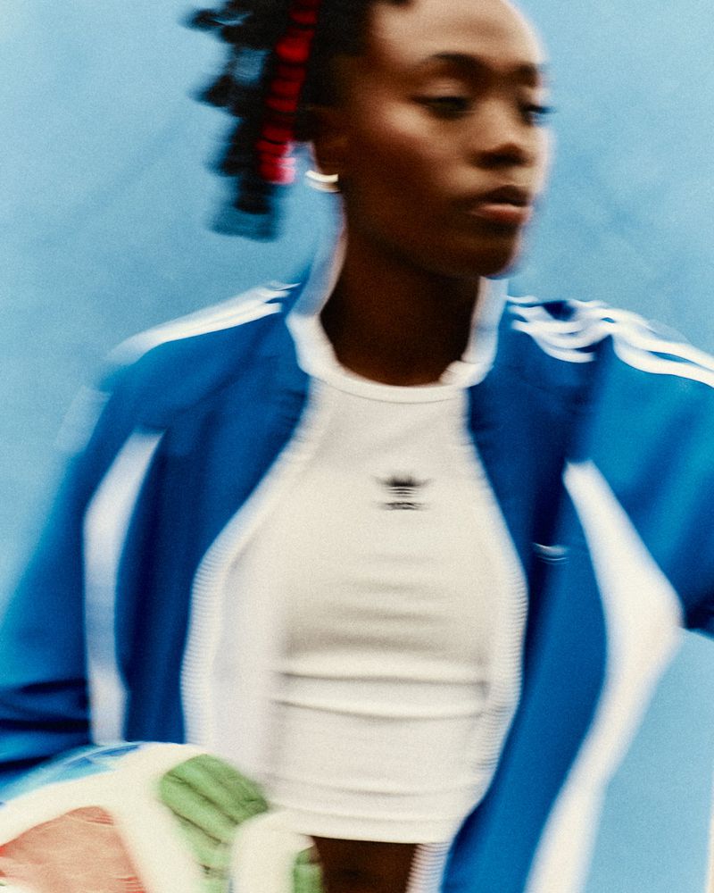 Athletic woman in blue track jacket and white top, blurred motion against blue backdrop, Adidas logo visible. Fashion, sportswear.