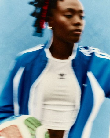 Athletic woman in blue track jacket and white top, blurred motion against blue backdrop, Adidas logo visible. Fashion, sportswear.