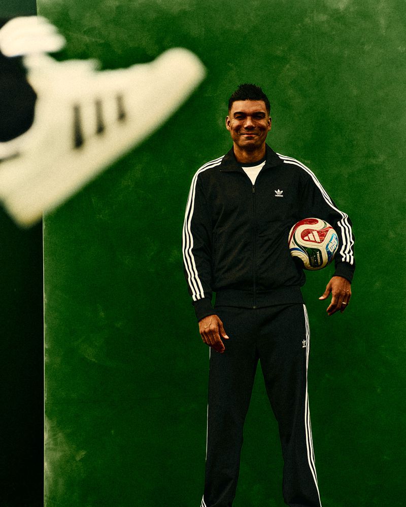 A full-shot of a man in a tracksuit holding a soccer ball, with a blurry shoe in the foreground against a green background.
