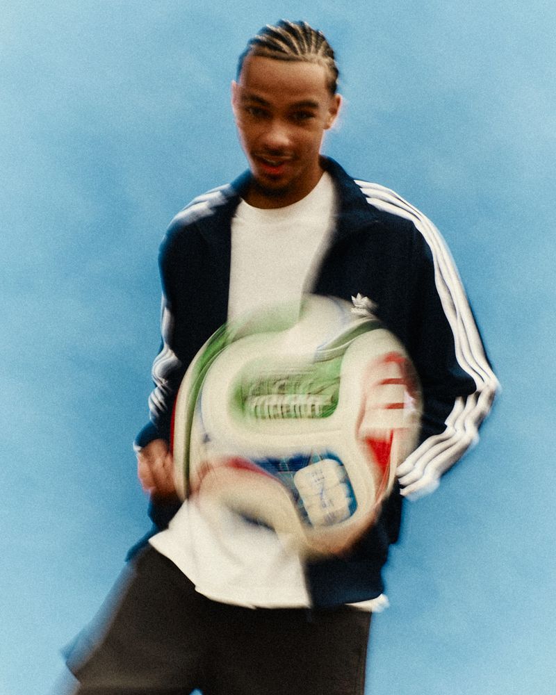 Young adult male poses with a soccer ball in front of a blue backdrop, wearing a tracksuit jacket and a white t-shirt.