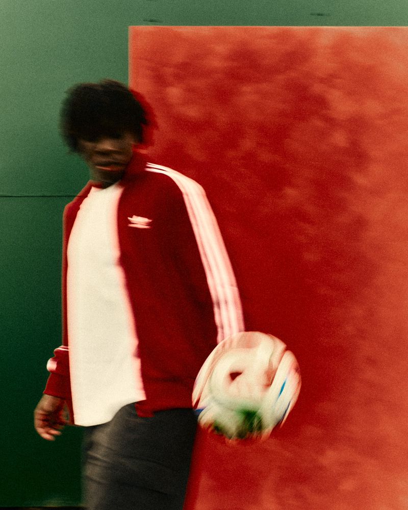 Man dribbling soccer ball wearing Adidas track jacket in motion, shot in front of green and red wall in an indoor setting.