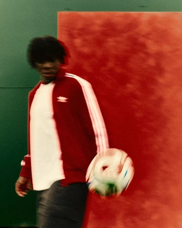 Man dribbling soccer ball wearing Adidas track jacket in motion, shot in front of green and red wall in an indoor setting.
