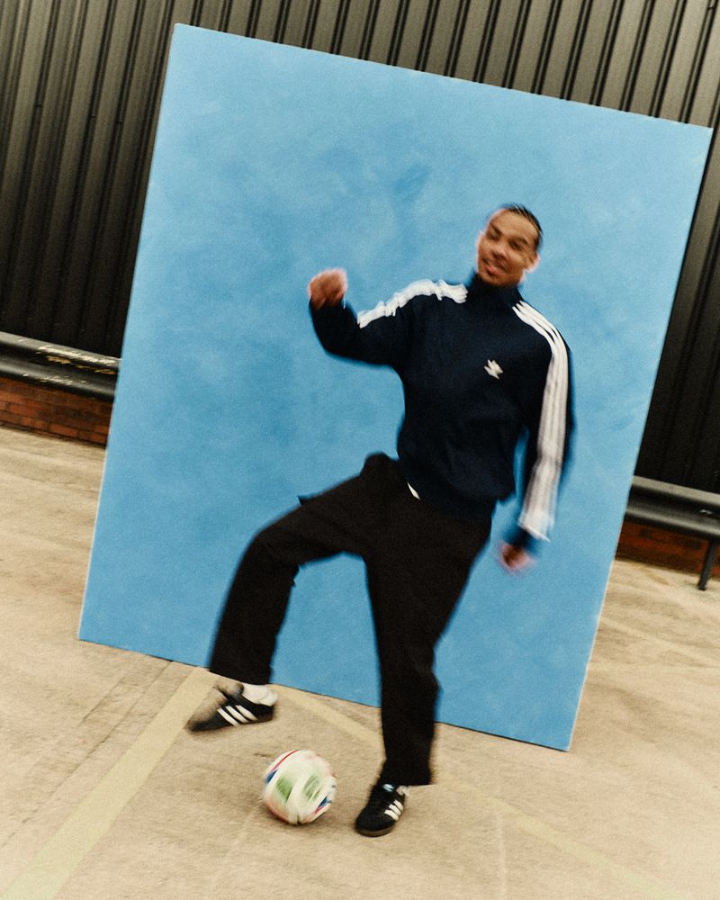 Man in Adidas tracksuit playing soccer with a soccer ball in front of a light blue screen in an indoor setting with overhead lighting.