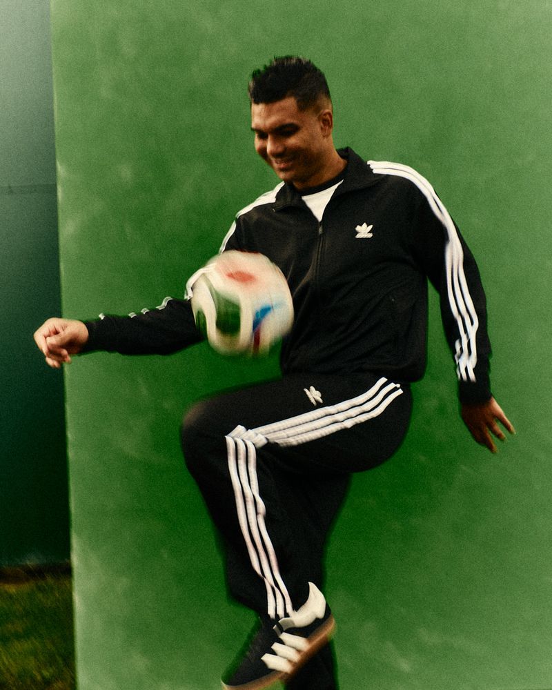Adult male playing soccer wearing a black Adidas track suit and black Adidas shoes standing outdoors with a green background.