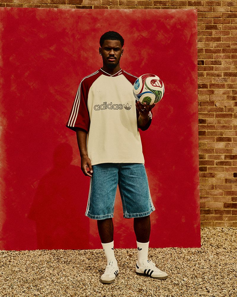 A young adult man stands holding a soccer ball in an outdoor setting, wearing a vintage-style Adidas t-shirt, denim shorts, socks, and sneakers.