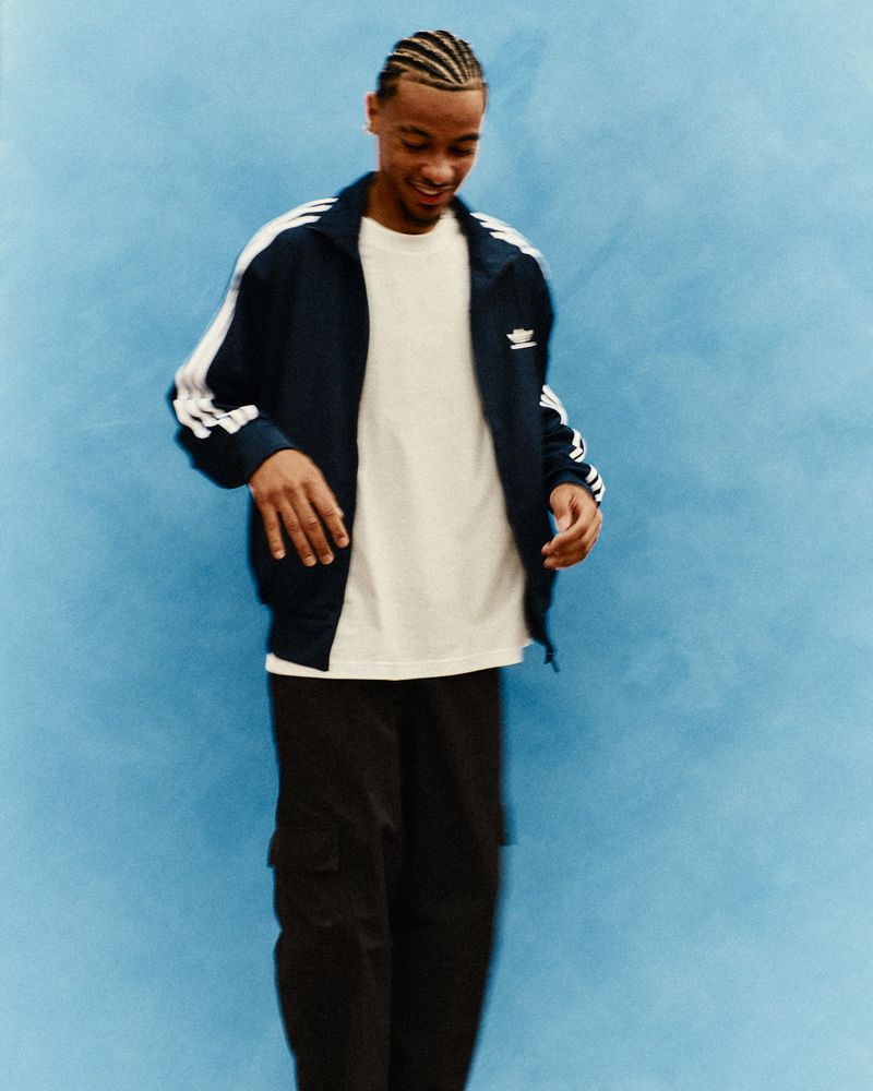 Young adult male wearing Adidas track jacket in a medium shot against a blue backdrop, smiling and posing for a fashion photograph.